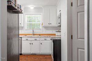 A small kitchen with white cabinets, a window with blinds above a sink, white subway tile backsplash, stainless steel dishwasher, microwave, toaster, and wood-look flooring. A white door is partially open on the right.