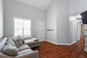 A living room with a gray sectional sofa, several patterned pillows, a window with blinds, a white front door, a wall-mounted coat rack, and wood flooring. A hallway leads to other rooms. Walls are light gray.