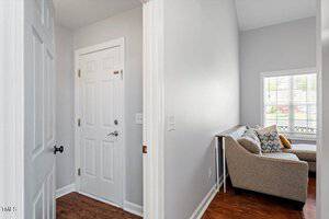 A small hallway with closed white doors and wood flooring leads to a cozy living area with a beige sectional sofa, colorful pillows, and a window letting in natural light.