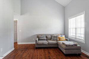 A gray sectional sofa with assorted pillows is placed in the corner of a room with light gray walls, a large window with blinds, and brown wood flooring. The room features high ceilings and simple decor.