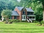 A two-story red brick house with white trim on 4139 Somerset Drive in Oxford features dark shutters, a welcoming front porch, and an American flag by the steps, all surrounded by green grass, shrubs, and mature trees on a sunny day.