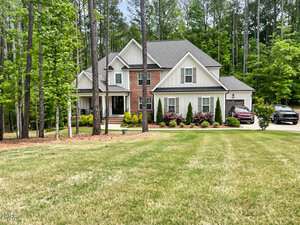 A two-story suburban house with brick and white siding at 1218 Rogers Farm Road, Wake Forest, is surrounded by tall trees. The front yard features trimmed grass and shrubs, with two cars parked in the driveway on the right side.