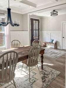 A dining room with a wooden table and six mismatched chairs, a dark chandelier, two rugs, light wood floors, white paneled walls, large windows with blinds, and a black front door letting in sunlight.