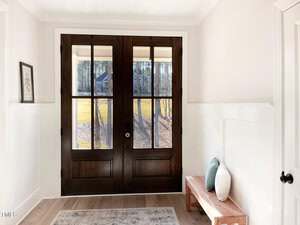A bright entryway with large double wooden doors featuring glass panels. A wooden bench with two throw pillows sits to the right, and a framed picture hangs on the left wall. Sunlight streams through the doors.