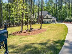 A two-story house with a front porch sits at the end of a curved driveway surrounded by trees and grass. A black mailbox numbered 218 is in the foreground, and a bicycle leans against a tree in the yard.