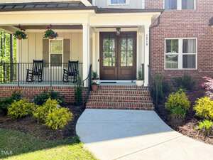 A house entrance with brick steps, a wooden double front door, two black rocking chairs on a covered porch, hanging flower baskets, potted plants, and landscaping with shrubs and bushes.