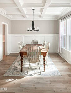 A dining room with a wooden table, six white chairs, a patterned rug, a black chandelier, wainscoted white walls, coffered ceiling, and large windows letting in natural light.