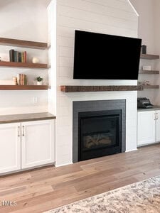 A modern living room with light wood floors, a white shiplap fireplace, a black-framed fireplace insert, a large mounted TV, built-in white cabinets, gray countertops, and wooden floating shelves with books and decor.