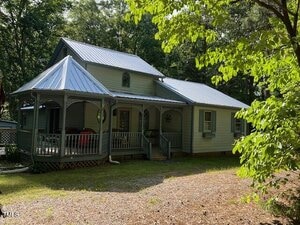 A light green house with a metal roof and a covered front porch sits at 358 Pool Rock Shores Lane, Henderson, surrounded by trees and a gravel driveway on a sunny day.