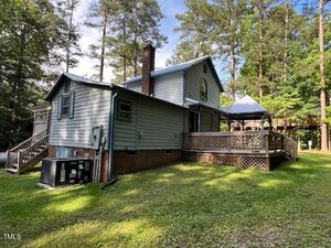 A two-story blue-gray house with a brick foundation, side deck, and gazebo is surrounded by tall trees and grass under a partly cloudy sky. Outdoor stairs lead to the upper level.