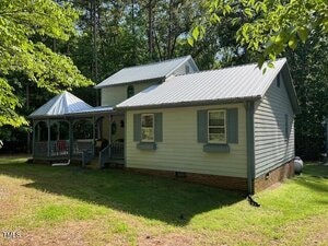 A small light gray house with a metal roof, two front-facing windows with blue shutters, a wooden porch with railing, and a gazebo attached, surrounded by green grass and trees.
