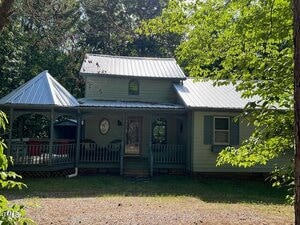 A light green house with a metal roof and a covered front porch sits among trees. The porch has a gazebo-style section on the left, two chairs near the entrance, and light blue shutters on the windows.