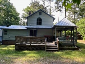A two-story house with light-colored siding, a metal roof, a wooden deck, and a covered gazebo area. The house is surrounded by trees and sits on a grassy lot.