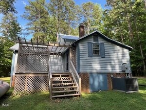 A small, light blue house with a brick foundation and a covered back porch. Wooden stairs lead up to the porch, which features a lattice railing and partial pergola. The house is surrounded by tall trees.