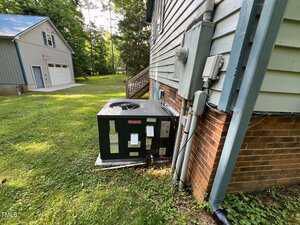 An outdoor HVAC unit is installed next to a brick house wall with utility meters and pipes attached. A detached garage and trees are visible in the background on a grassy lawn.