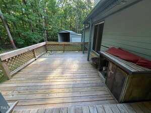 A wooden deck attached to a light green house, with a lattice railing, covered grill, and a small metal shed in the background. The deck overlooks a wooded area.