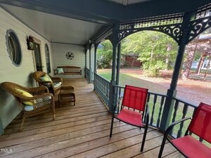 A covered wooden porch with wicker chairs, a wooden bench, red chairs, and decorative railings, overlooking a yard with grass and trees in the background.