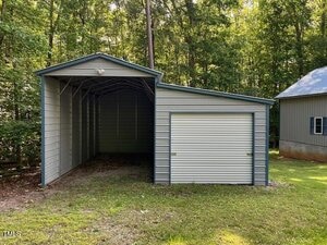 A metal carport with an attached garage featuring a roll-up door is situated on a grassy area near a wooded lot. Another building with a metal roof is partially visible on the right.