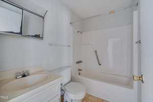 A small bathroom with a white vanity and sink, a toilet, and a bathtub-shower combination with a grab bar. The floor has beige tiles and there is a wall-mounted mirror above the sink.
