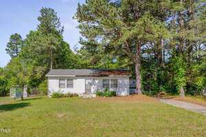 A small white single-story house with a gray roof sits at 1109 N Pinkston St. Street, Henderson, surrounded by tall pine trees and a grassy yard. A paved driveway lies to the right and a wooden fence is partially visible in the background.