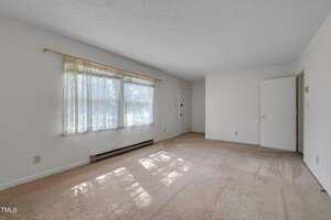 An empty room with beige carpet, white walls, a large window with sheer curtains, a baseboard heater below the window, and an open white door leading to another area. Natural light fills the room.