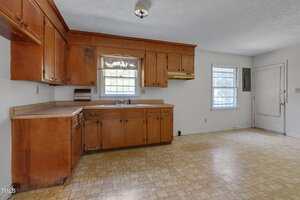 A kitchen with wooden cabinets, a double sink under a window, beige countertops, worn linoleum flooring, and two windows allowing natural light. The space appears empty and extends to an entry door on the right.
