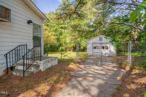 A concrete walkway leads to a metal gate in a chain-link fence, separating a small house with steps and railing on the left from a detached white garage surrounded by trees and greenery in the background.