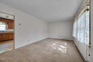 A mostly empty living room with beige carpet, white walls, and a large window with lace curtains. The room connects to a kitchen with wooden cabinets and tiled flooring visible through an open doorway.