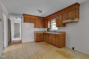 Empty kitchen with wooden upper and lower cabinets, beige countertops, a window above the sink, tile flooring, and white walls. There is an empty space for an appliance and a hallway visible in the background.
