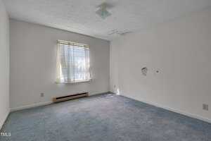 Empty room with white walls, a window with blinds letting in sunlight, blue carpeted floor, a heater under the window, a ceiling light fixture, water stains on the ceiling, and a hole in the right wall.