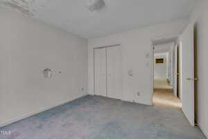 A small empty room with white walls, two holes in the wall, a white closet with double doors, a light fixture on the ceiling, and worn blue carpet. An open door leads to a hallway with beige carpeting.