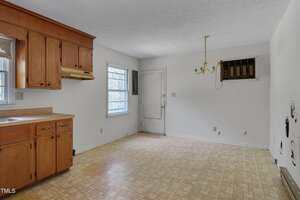 An empty kitchen with wooden cabinets, a beige countertop, linoleum flooring, a window, a wall-mounted air conditioning unit, a ceiling light fixture missing shades, and exposed plumbing on one wall.