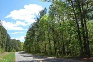 A two-lane road curves to the left on Egypt Moutain Road in Kittrell, bordered by tall green trees under a partly cloudy blue sky. Sunlight filters through the foliage, and the road appears empty.