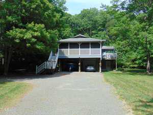 Nestled in Kittrell at 4144 Fairport Road, this raised house with a large screened porch sits above a three-car carport. Two vehicles are parked underneath, and a gravel driveway winds through the treed property.