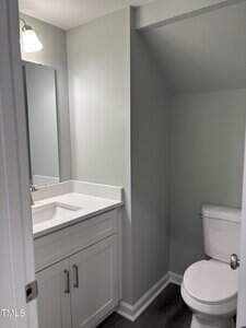 A small bathroom with light gray walls, a white vanity with a sink and countertop, a rectangular mirror above, and a toilet positioned under a sloped ceiling. The floor is dark wood or laminate.