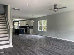 Open-concept living area with gray flooring, light gray walls, large windows, a staircase on the left, and a modern kitchen with white cabinets, a dark island, and stainless steel appliances in the background.