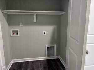 A laundry room with light gray walls, white trim, dark wood flooring, plumbing hookups for a washer and dryer, an electrical outlet, and a white shelf mounted on the wall.