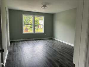 An empty room with gray walls, dark wood-style flooring, a ceiling light fixture, and a large window showing a view of trees and a house outside. The room appears clean and well-lit.