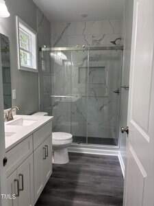 A modern bathroom with white cabinets, a white countertop with dual sinks, a toilet, and a glass-enclosed shower with marble-patterned walls and dark tile flooring. A small window lets in natural light.