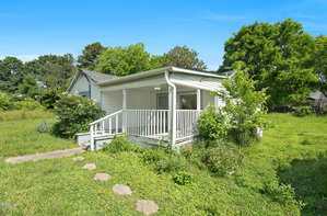 A small white house with a covered front porch and railing, surrounded by overgrown grass and bushes. There is a concrete path leading to the porch, and trees are visible in the background under a clear blue sky.