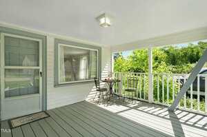 A covered porch with gray flooring, white railings, a gray-trimmed door, a large window, and a small black metal table with two matching chairs. Sunlight filters in and greenery is visible beyond the railing.
