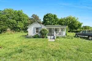 A small white single-story house at 441 Cedar Street, Henderson, features a covered front porch and sits on a large grassy lot surrounded by trees. A black pickup truck is parked to the right under a clear blue sky.