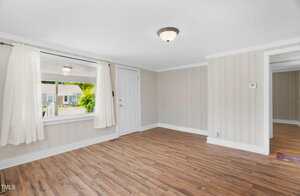 A small, empty room with light-colored wood flooring, beige striped walls, a white ceiling, a single ceiling light, a window with white curtains, and a white door leading outside.