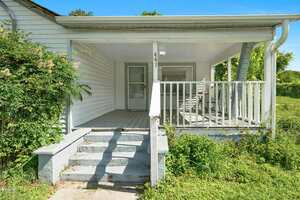 Front view of a white house with a covered porch, steps leading up, a railing, and overgrown greenery on either side. The house has two visible doors and a light fixture by the entrance.