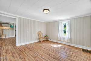 A bright room with light wood floors, beige vertical panel walls, a white ceiling, a window with sheer curtains, a wooden chair, and a small table. An open doorway reveals a connected kitchen area.