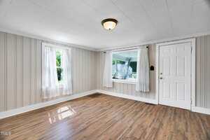 Empty room with light-colored wood flooring, two windows with sheer white curtains, beige vertical wall paneling, a white ceiling with a single light fixture, and a white front door.