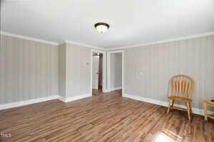 A mostly empty room with light wood flooring, gray vertical panel walls, white baseboards and trim, a ceiling light, a single wooden chair, and a small table near a doorway leading to another room.