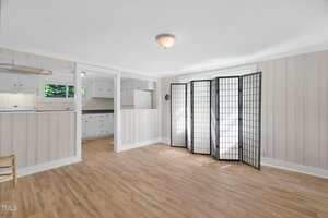 A bright room with light wood flooring, striped wallpaper, a ceiling light, a folding room divider with white panels, and an open view into a white kitchen with cabinetry and a window.