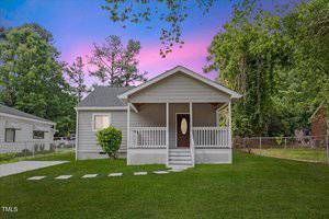 A small gray house at 128 W Parker Street in Henderson features a covered front porch, white trim, and a dark front door. Stepping stones lead across the grassy lawn, with trees and neighboring houses silhouetted under a purple sky.