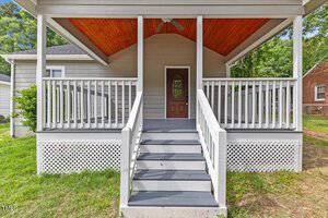 A front porch with gray steps and white railings leads to a wooden door. The porch has a vaulted ceiling with exposed wood, and white lattice skirting surrounds the base. Grass and trees are visible in the background.